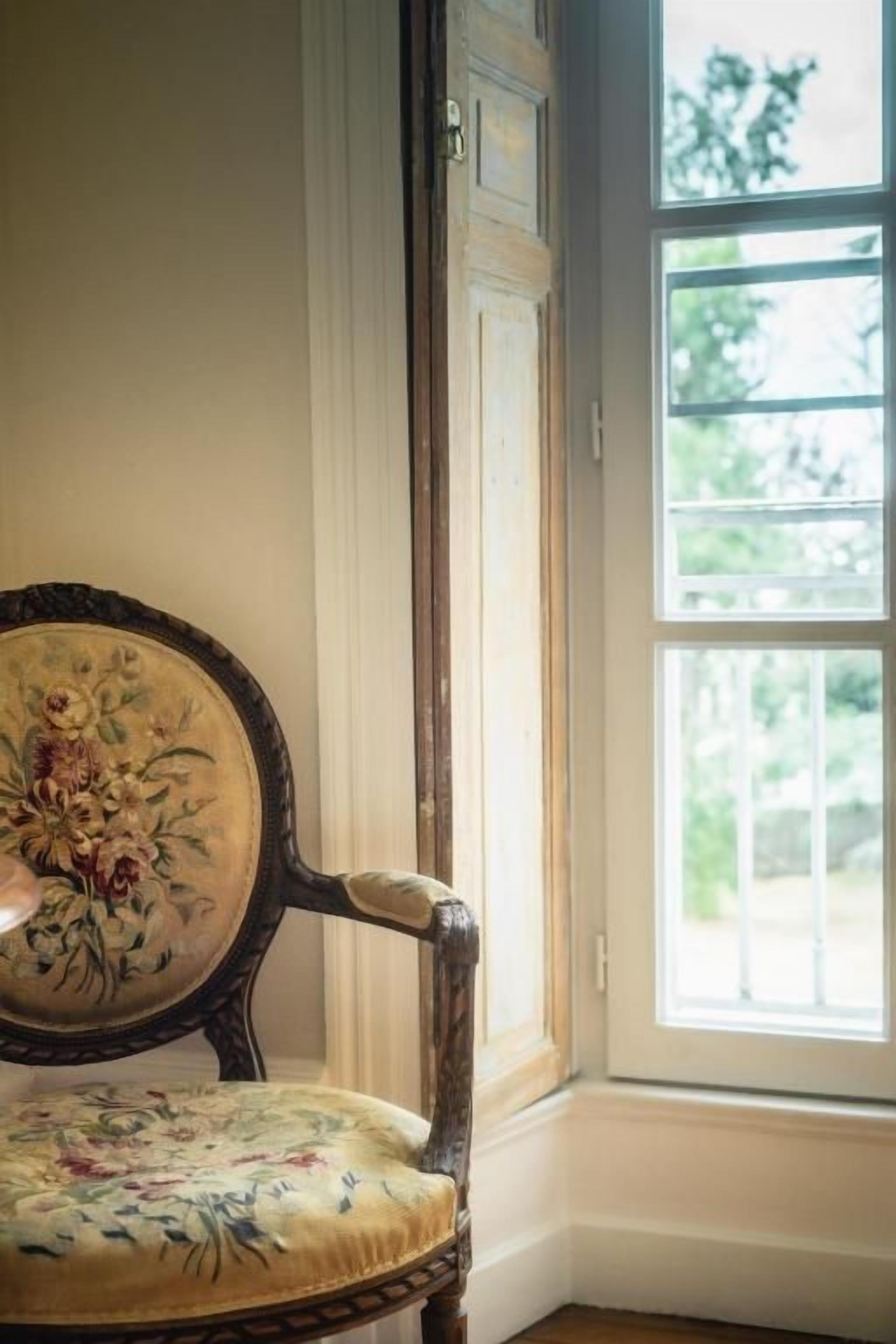 A corner of a room with a wooden chair with floral upholstery and a window with a view of greenery.