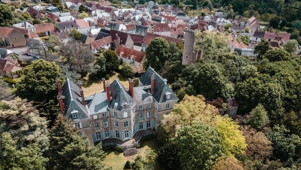 A large, historic building with multiple chimneys, surrounded by a dense forest and other residential buildings.