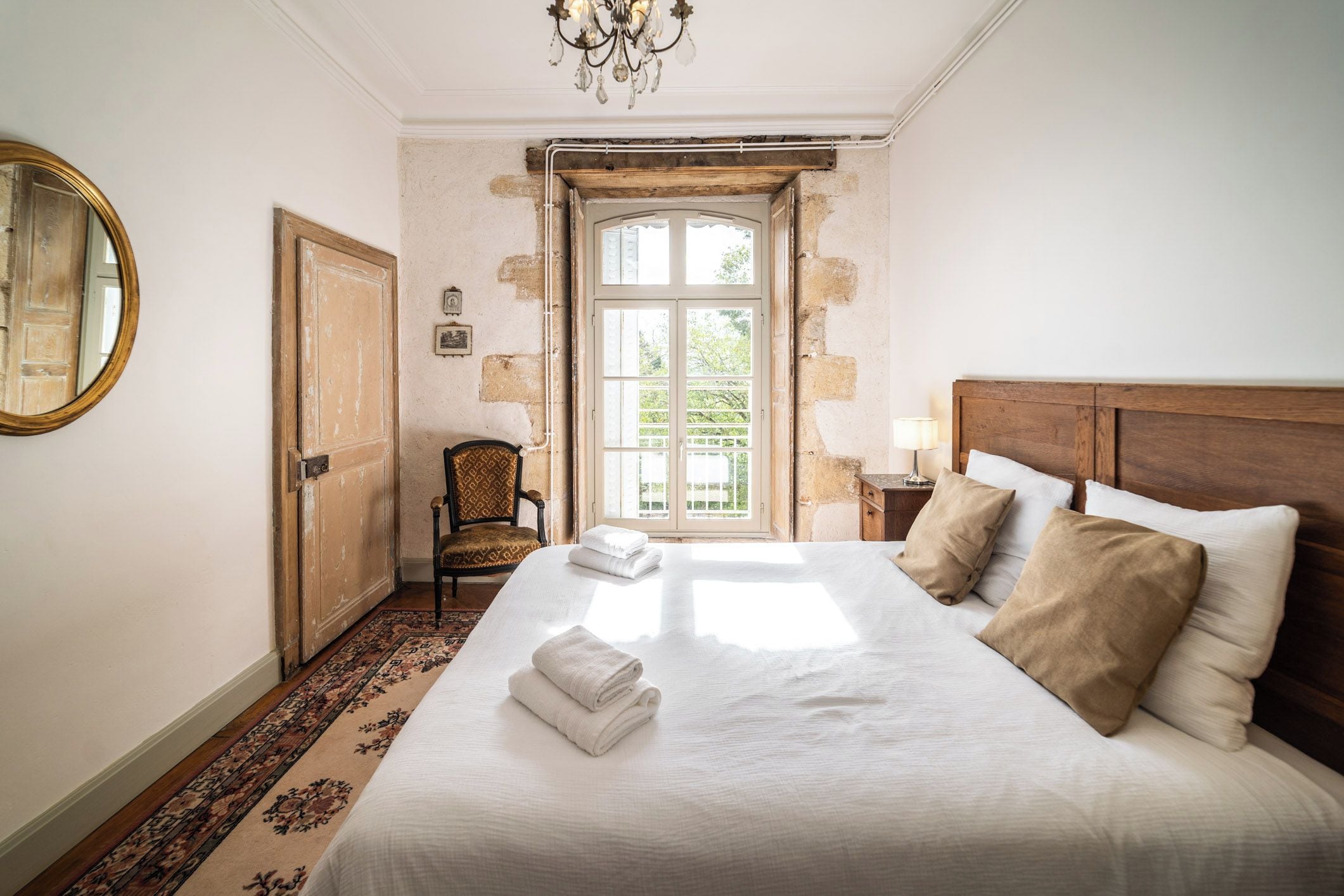 Un dormitorio con una cama grande, cabecera de madera, una araña, una silla y vistas por una ventana.