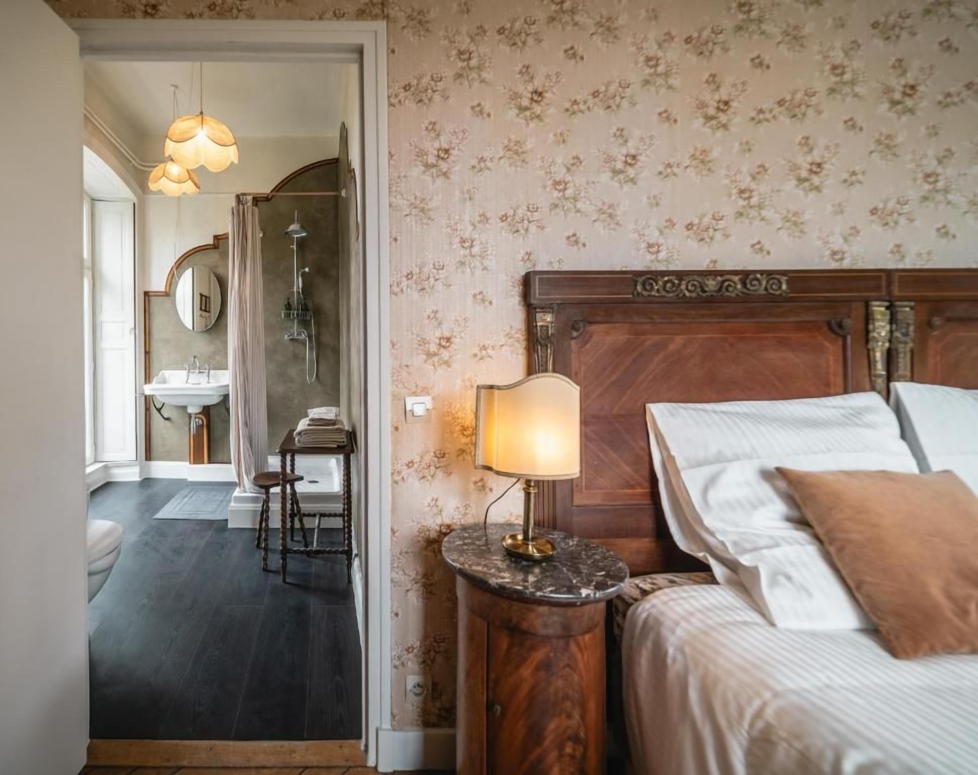 Een hotelkamer met een houten hoofdbord, een bedlampje, een bed met witte lakens, een nachtkastje en uitzicht op de badkamer.