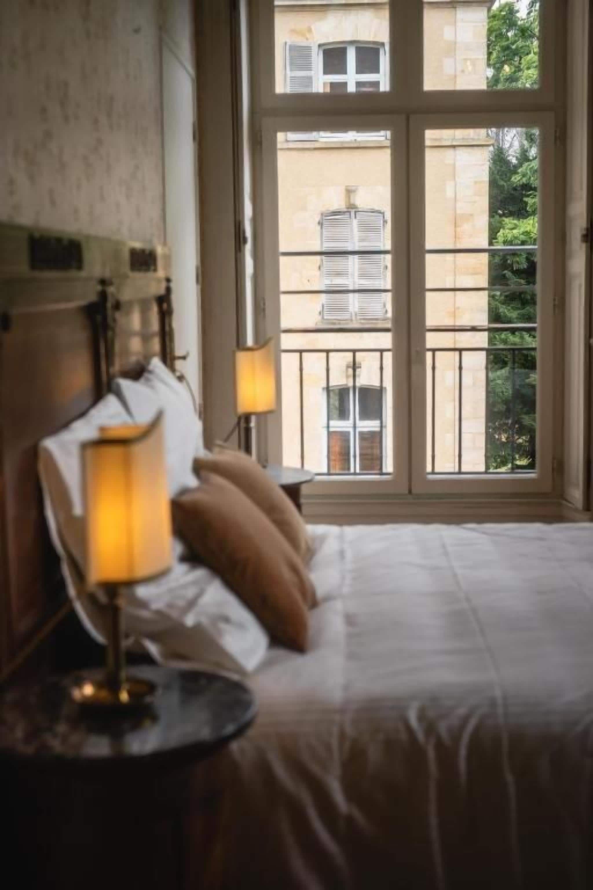 A hotel room with a bed, a nightstand with a lamp, and a window with a view of buildings and greenery.