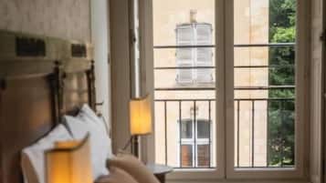 A hotel room with a bed, a nightstand with a lamp, and a window with a view of buildings and greenery.