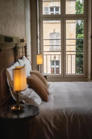 A hotel room with a bed, a nightstand with a lamp, and a window with a view of buildings and greenery. - Château de Sainte-Sévère (Sainte-Sévère-sur-Indre)