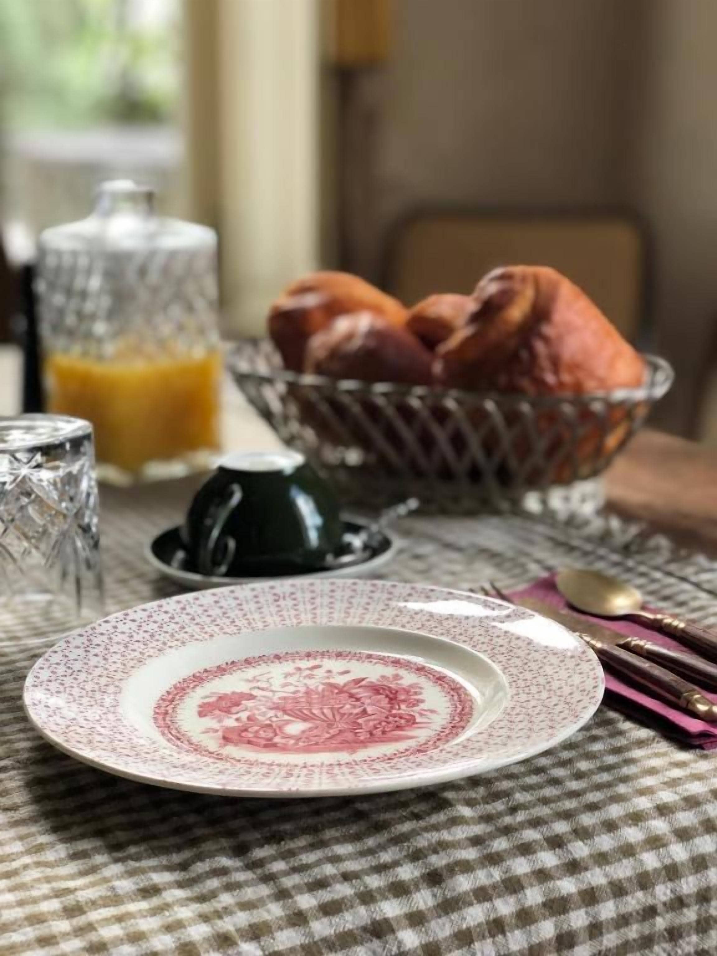 A dining table with a checkered tablecloth, a plate with a floral design, a basket of bread, a glass container, and a teapot.