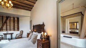 A bedroom with a large bed, wooden headboard, bedside tables with lamps, a chandelier, and a view of the bathroom through an open door.