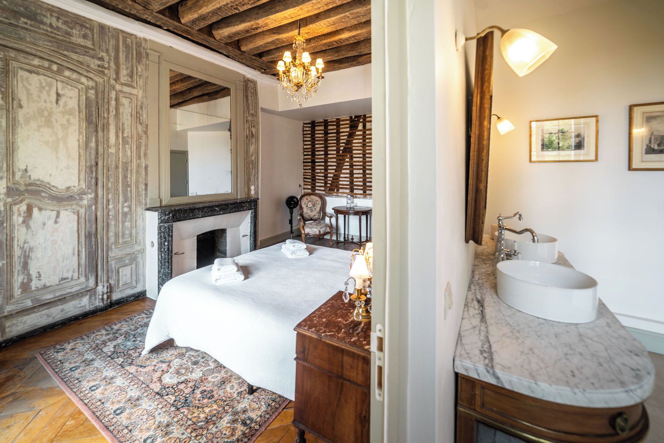 A bedroom with a large bed, a fireplace, a wooden wardrobe, and a bathroom with a sink and mirror.