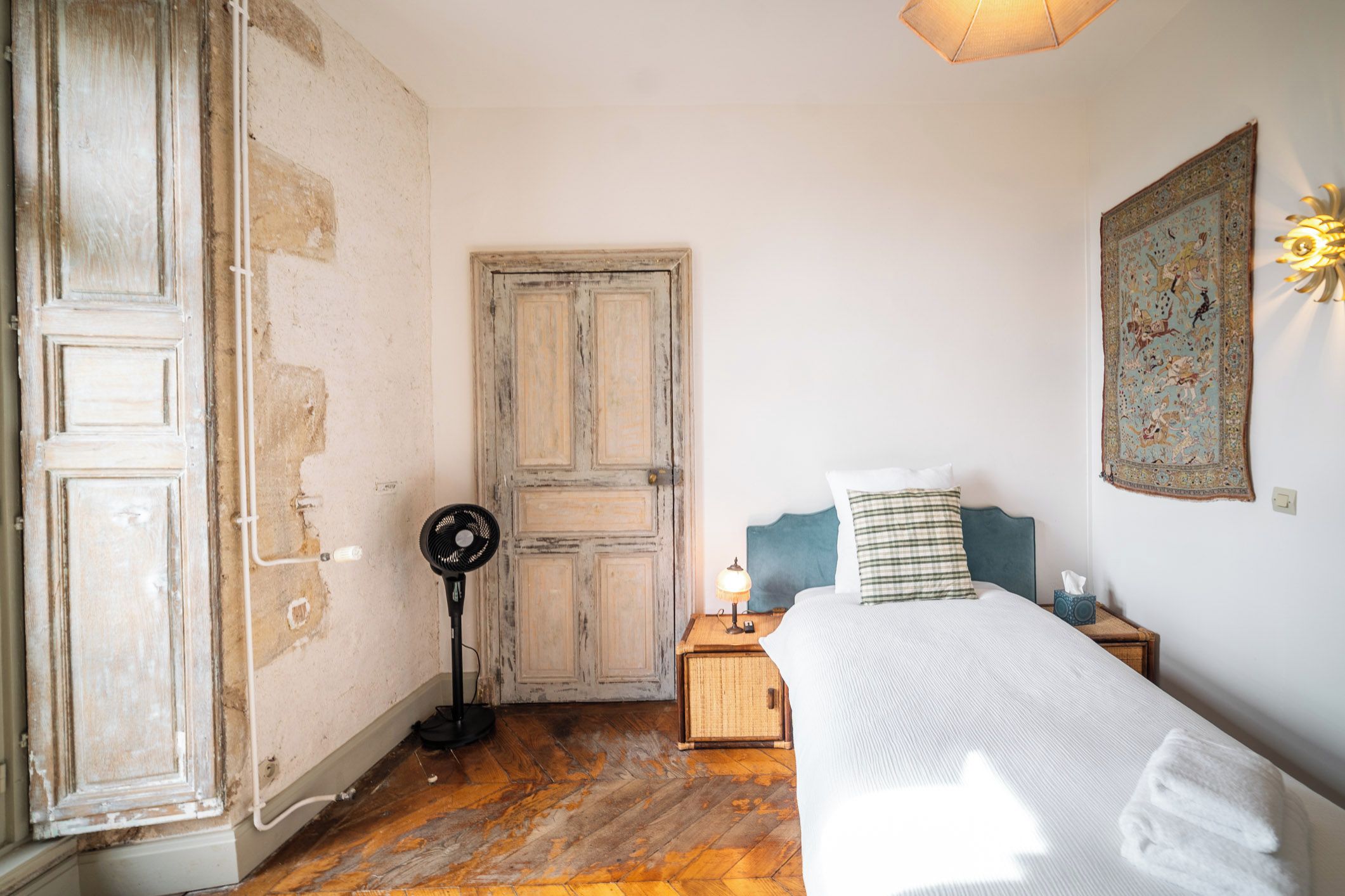 Quarto com piso de madeira, cama com cobertura branca, mesa de cabeceira com abajur e tapeçaria na parede.
