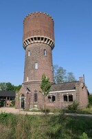 Una torre de ladrillo con base cilíndrica y un extremo superior cilíndrico más pequeño, rodeada por un edificio con ventanas de arco y una puerta.