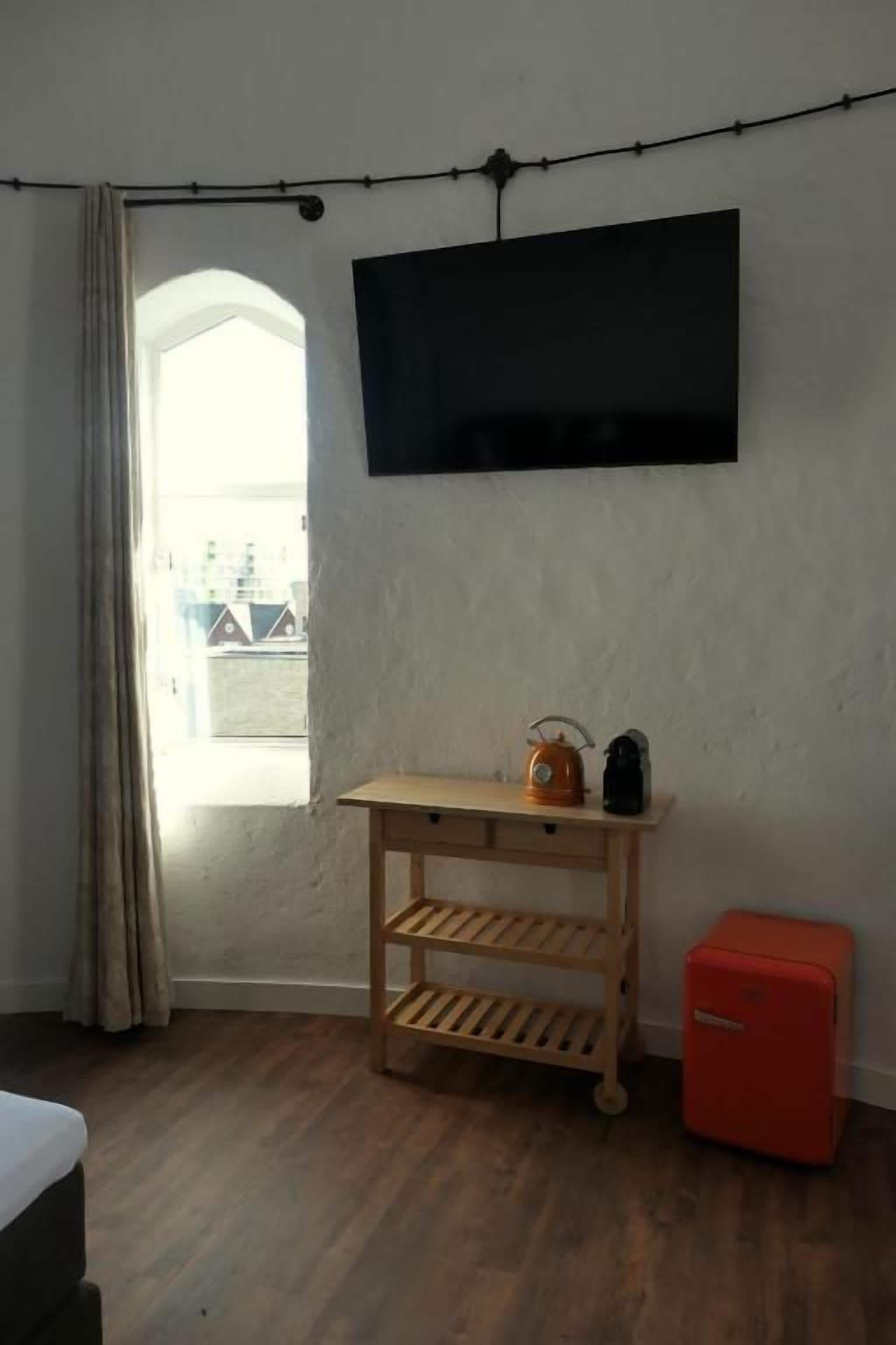 Una habitación con piso de madera, un televisor instalado en la pared, un mueble pequeño rojo y una ventana con cortinas.