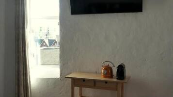 Una habitación con piso de madera, un televisor instalado en la pared, un mueble pequeño rojo y una ventana con cortinas.