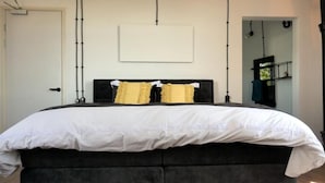 Una cama con cabecera negra y colcha blanca, dos almohadas y una lámpara fijada a la pared.