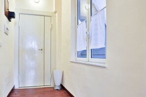 Ein Flur mit einer geschlossenen Tür, einem Fenster mit Vorhängen und einem Mülleimer.