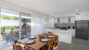 A modern kitchen with a central island, a dining area with a table set for two, and a view of the outdoors through large sliding glass doors. - Kiama Hamptons Hideaway - Spa & Sauna Retreat (Kiama)