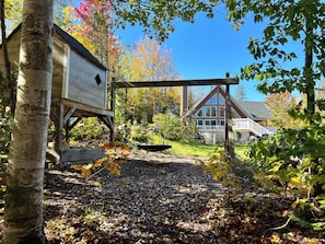 A wooden swing set, a birdhouse, and a house with a deck in a garden with autumn leaves. - 4-Bedroom Chalet with Spa and Pool in Rawdon (Rawdon)