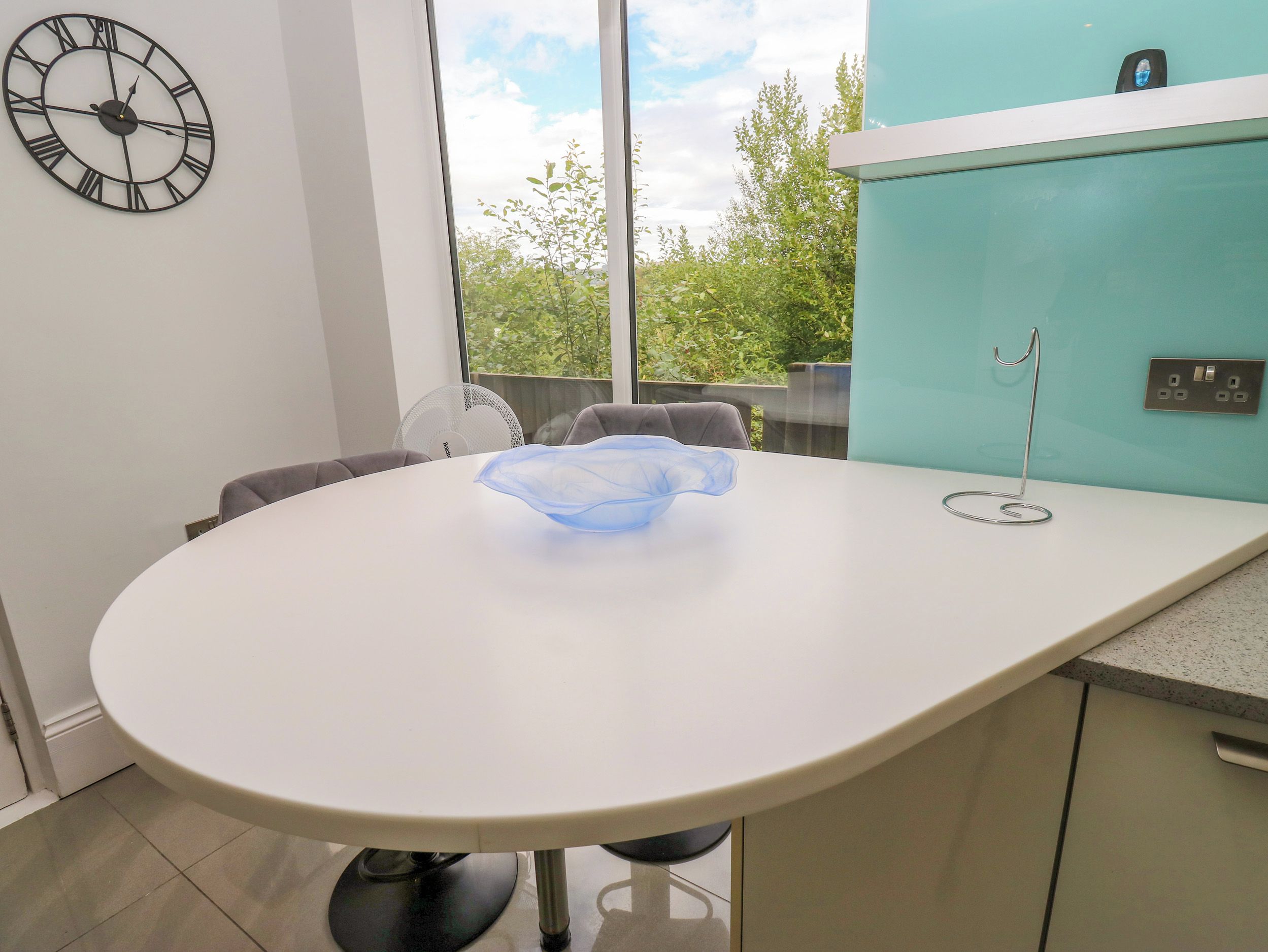 A modern dining area with a round table, chairs, a wall clock, and a view of greenery outside.