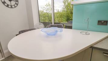 A modern dining area with a round table, chairs, a wall clock, and a view of greenery outside.