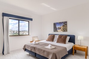 A hotel room with a bed, a bedside table, a lamp, a painting on the wall, and a window with curtains. - #425 Cozy Getaway in Albufeira | Shared Pools (Albufeira)