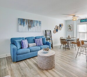 Ein Wohnzimmer mit einem blauen Sofa, einem runden Korbtisch, einem Esstisch mit Korbstühlen, einem Deckenventilator und einem an der Wand befestigten abstrakten Gemälde.