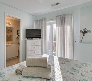 Une chambre à coucher avec un lit, un téléviseur posé sur un meuble, une salle de bain avec un lavabo et un miroir, et une vue sur l’extérieur.