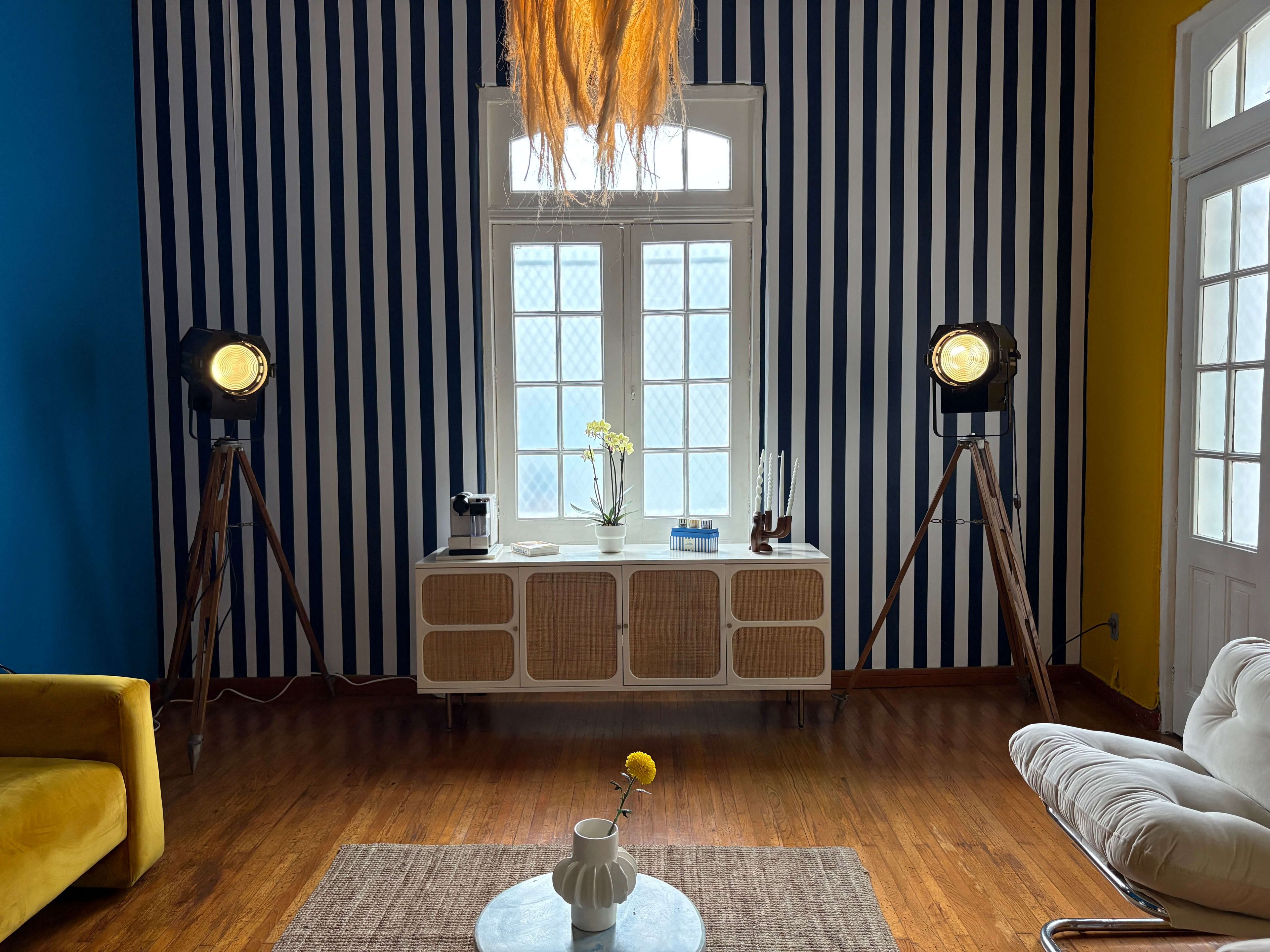A room with striped wallpaper, a wooden floor, a white sofa, a yellow armchair, a small table with a vase, and two floor lamps.