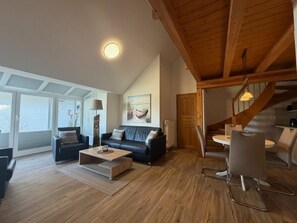 Ein modernes Wohnzimmer mit Holzbalkendecke, einem schwarzen Sofa, einem Couchtisch und einem Essbereich mit Stühlen.
