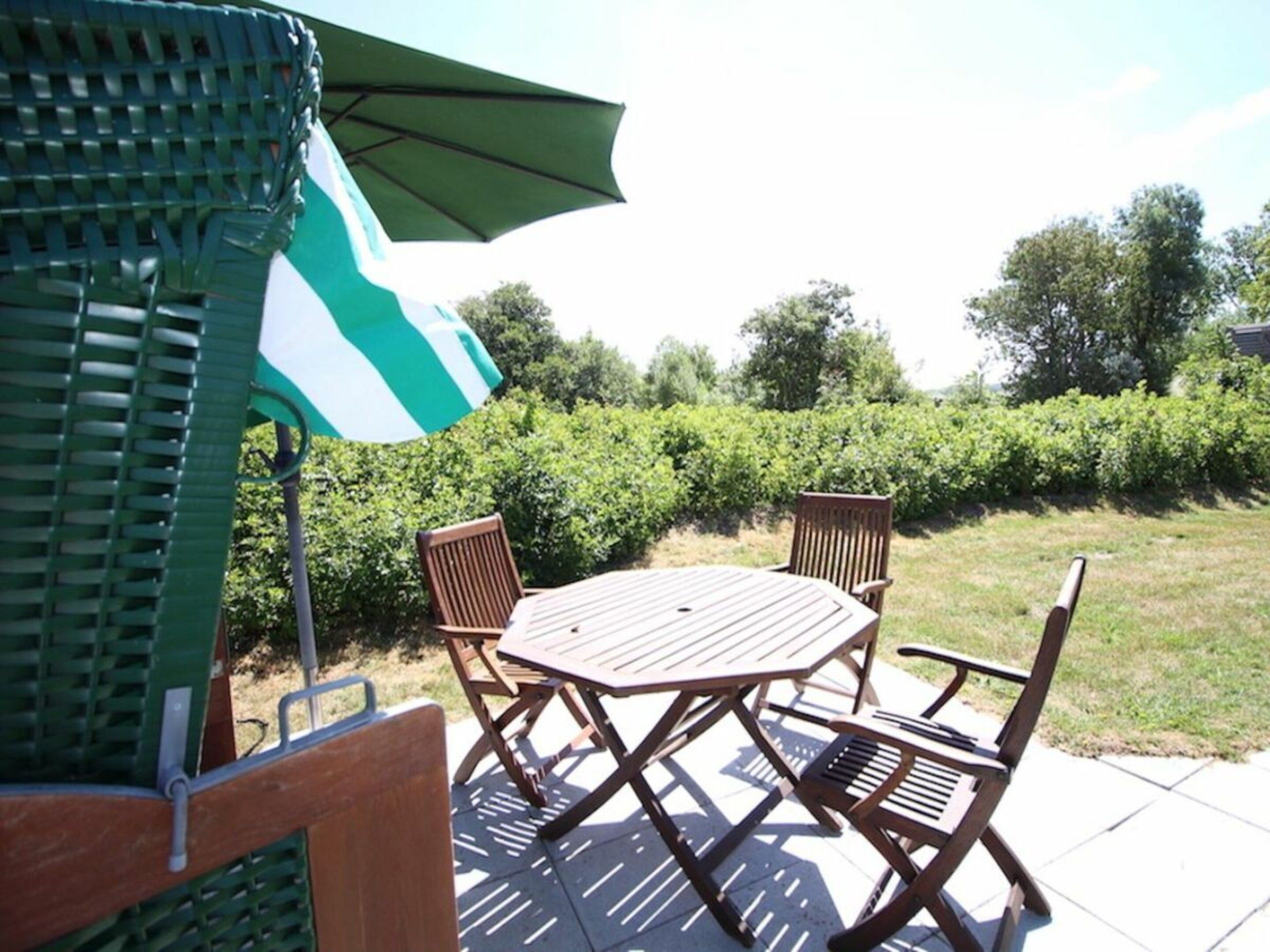 An outdoor patio with a table and chairs, a green umbrella, and a hedge in the background.