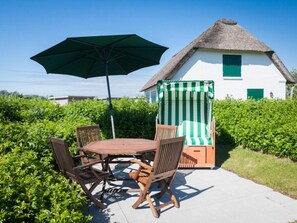 Een terras met een houten tafel en stoelen, een groene parasol en een rieten gebouw met een gestreepte strandstoel.