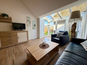 Ein modernes Wohnzimmer mit Couch, Holz-Couchtisch, Fernseher auf einer Holzkommode und einem großen Fenster mit Blick ins Freie.