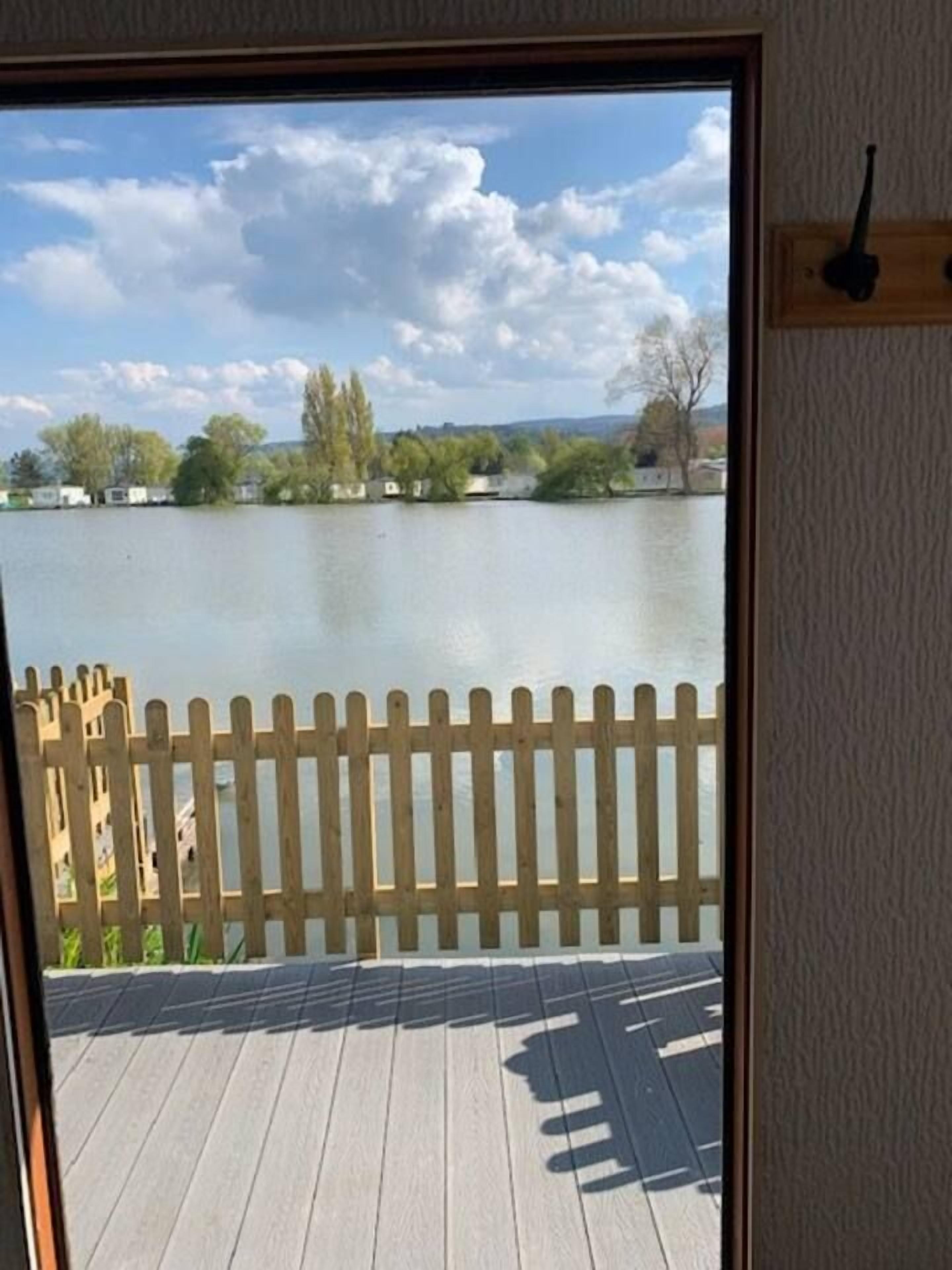 Vista a través de una ventana con terraza y cerca de madera, con vista a un cuerpo de agua.