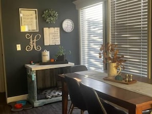 Un comedor con una mesa, sillas, una cama para mascotas, una planta y una ventana con persianas.