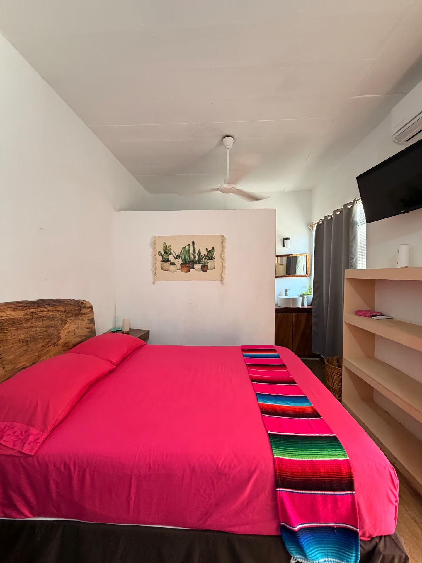 Un dormitorio con cama, ventilador de techo, televisión montada en la pared, baño visible a través de una puerta abierta y una obra de arte en la pared.