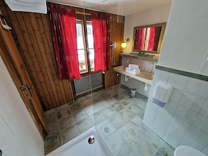 A bathroom with a wooden wall, a window with red curtains, a sink, a mirror, and a bathtub. - Schlosswirt (Grosskirchheim)