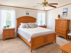 Une chambre à coucher avec un lit en bois, une table de chevet, une commode, un ventilateur de plafond et une fenêtre avec des stores.