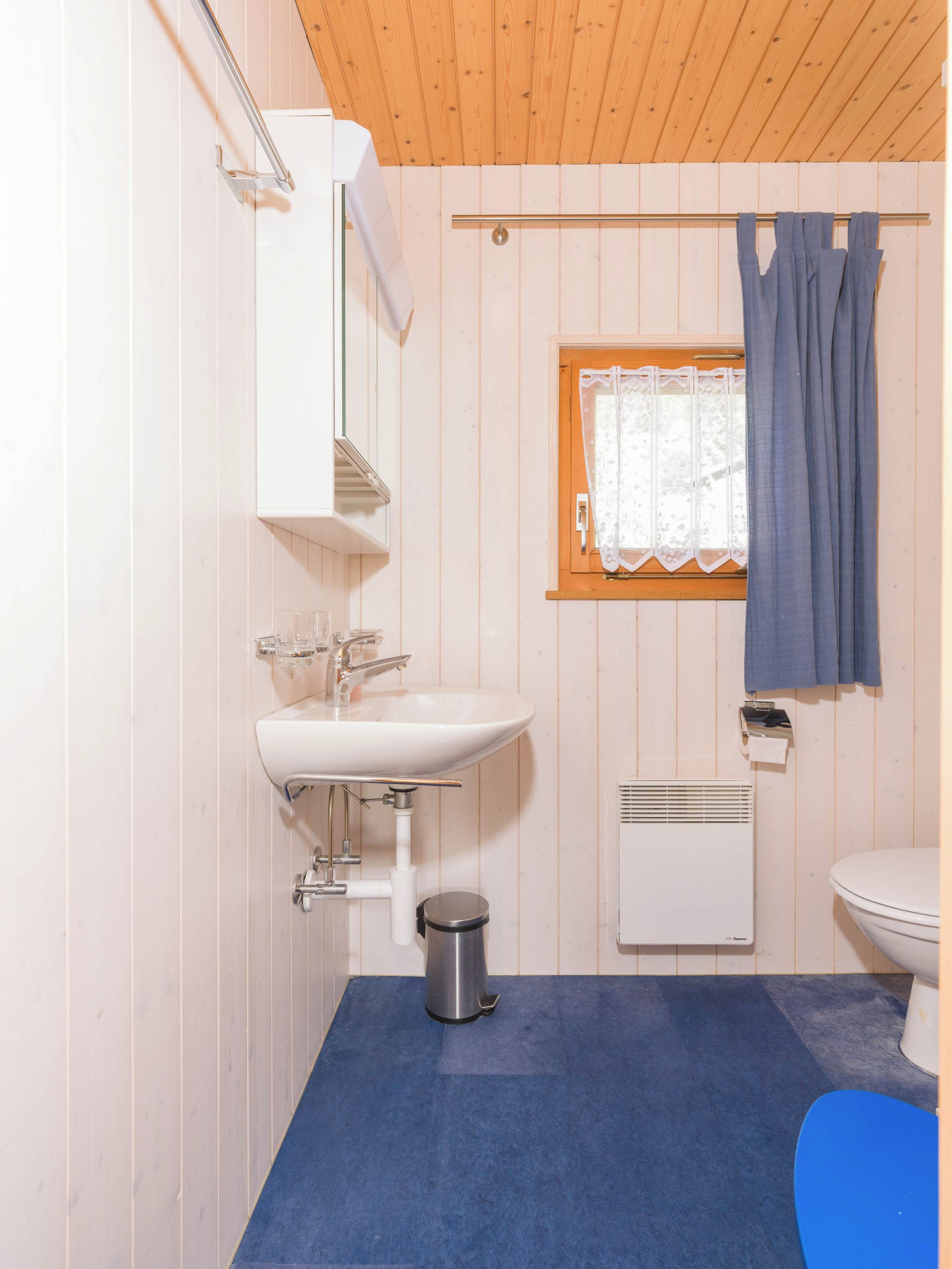 A small bathroom with a white sink, a toilet, a window with a curtain, and a blue floor.