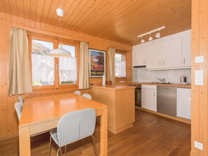 En inredning med träpanel, ett matbord, stolar, ett kök med vita skåp och ett fönster med gardiner.