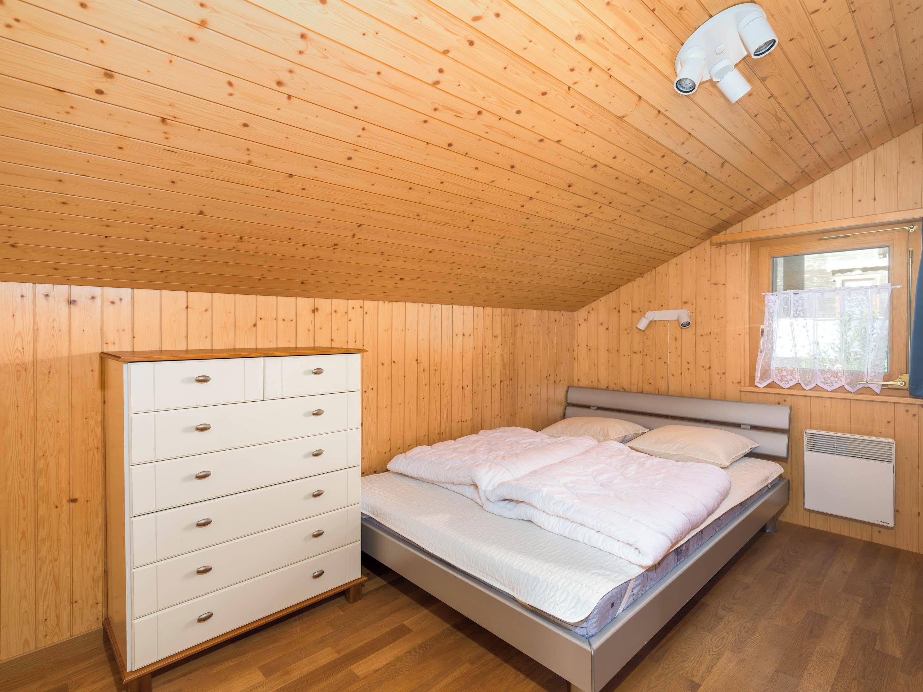 A bedroom with a wooden ceiling, a bed with white bedding, a white dresser with drawers, and a window with a curtain.