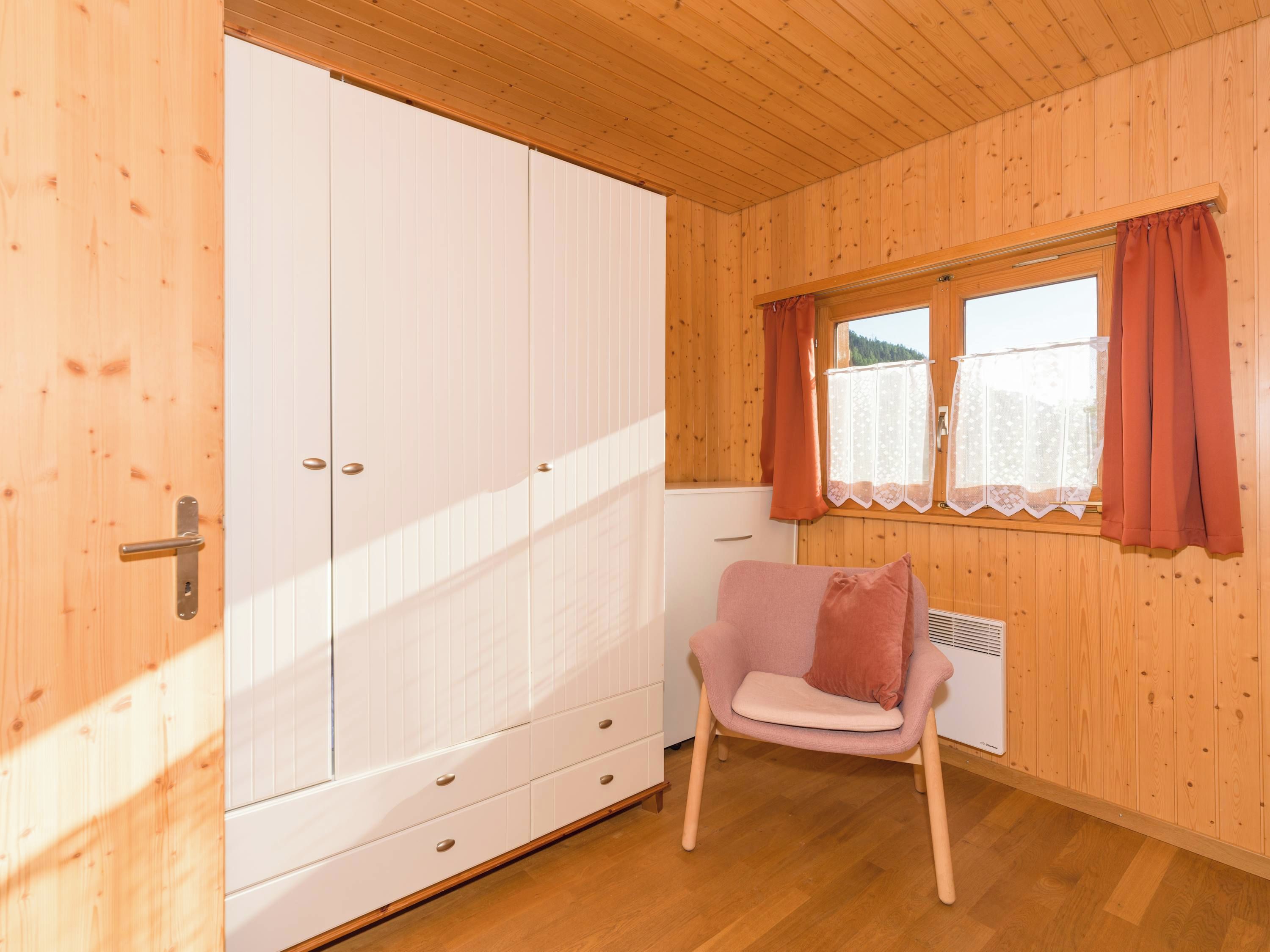 Une pièce aux murs en bois, avec une armoire blanche, une chaise rose et une fenêtre avec des rideaux.