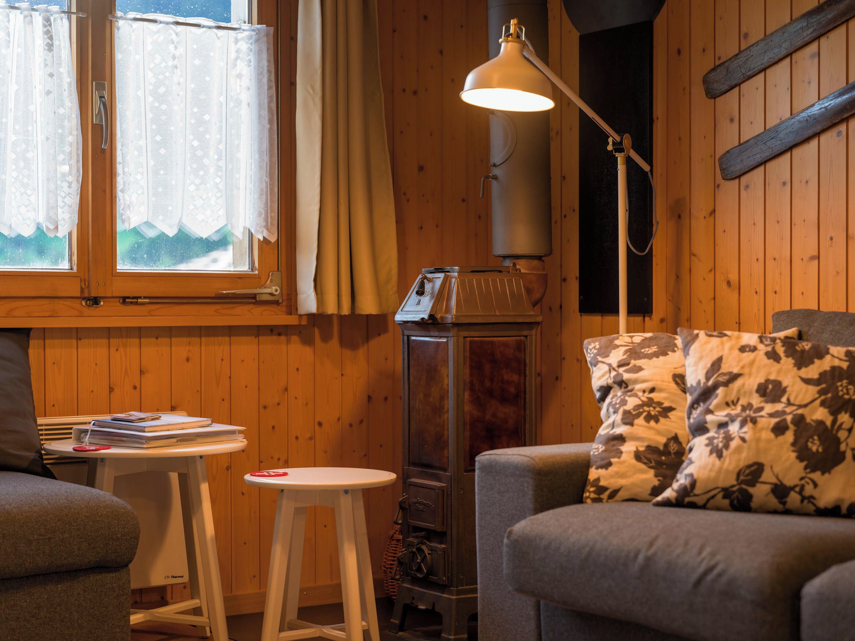 A cozy room with wooden walls, a sofa with floral cushions, a small white table with a book and a red object, and a tall lamp.