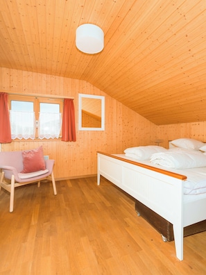Un dormitorio con techo de madera, una cama con ropa blanca, una silla con cojín y una ventana con cortinas.
