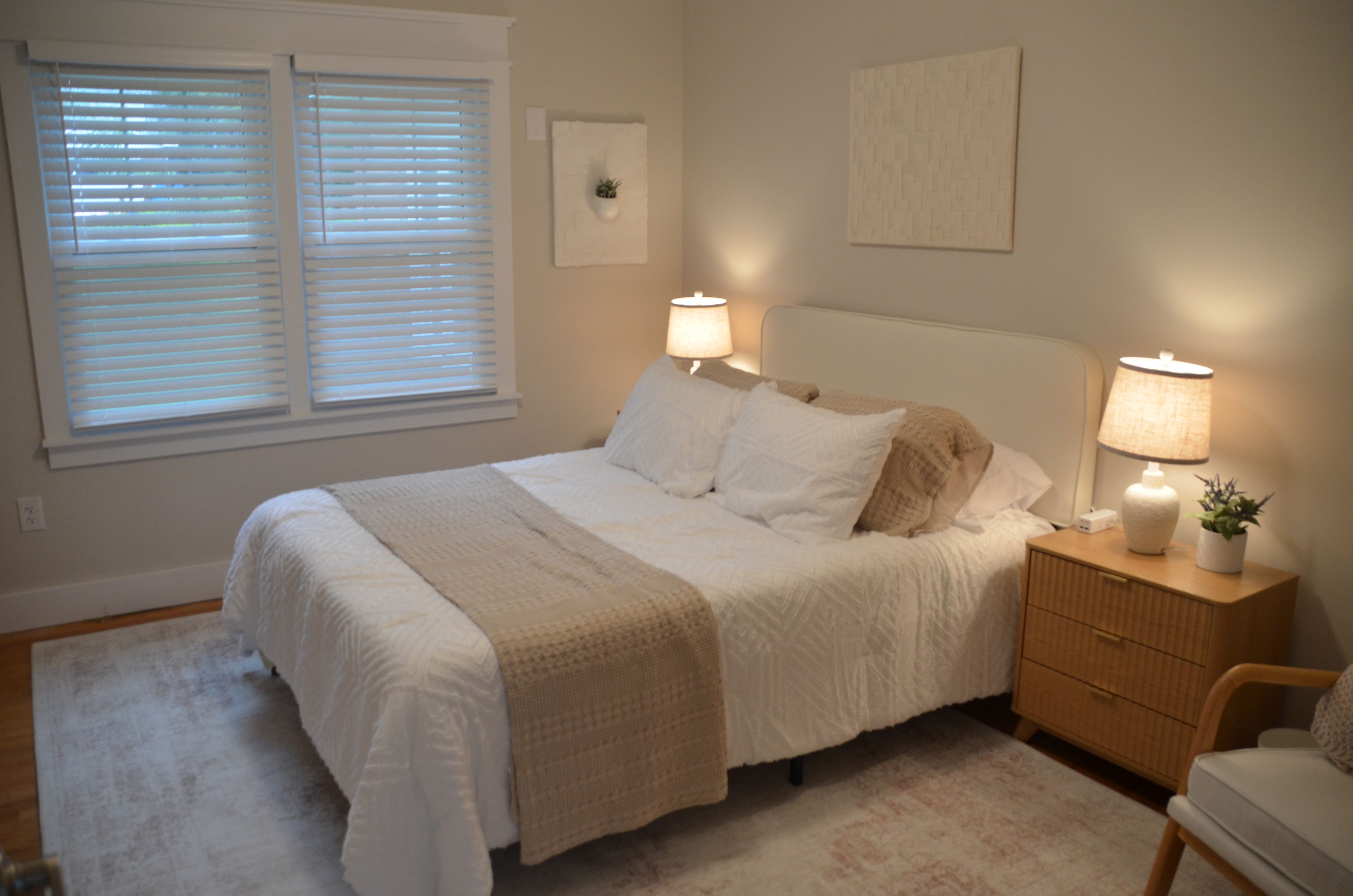 A bedroom with a bed, bedside lamps, a nightstand, a chair, and a window with blinds.