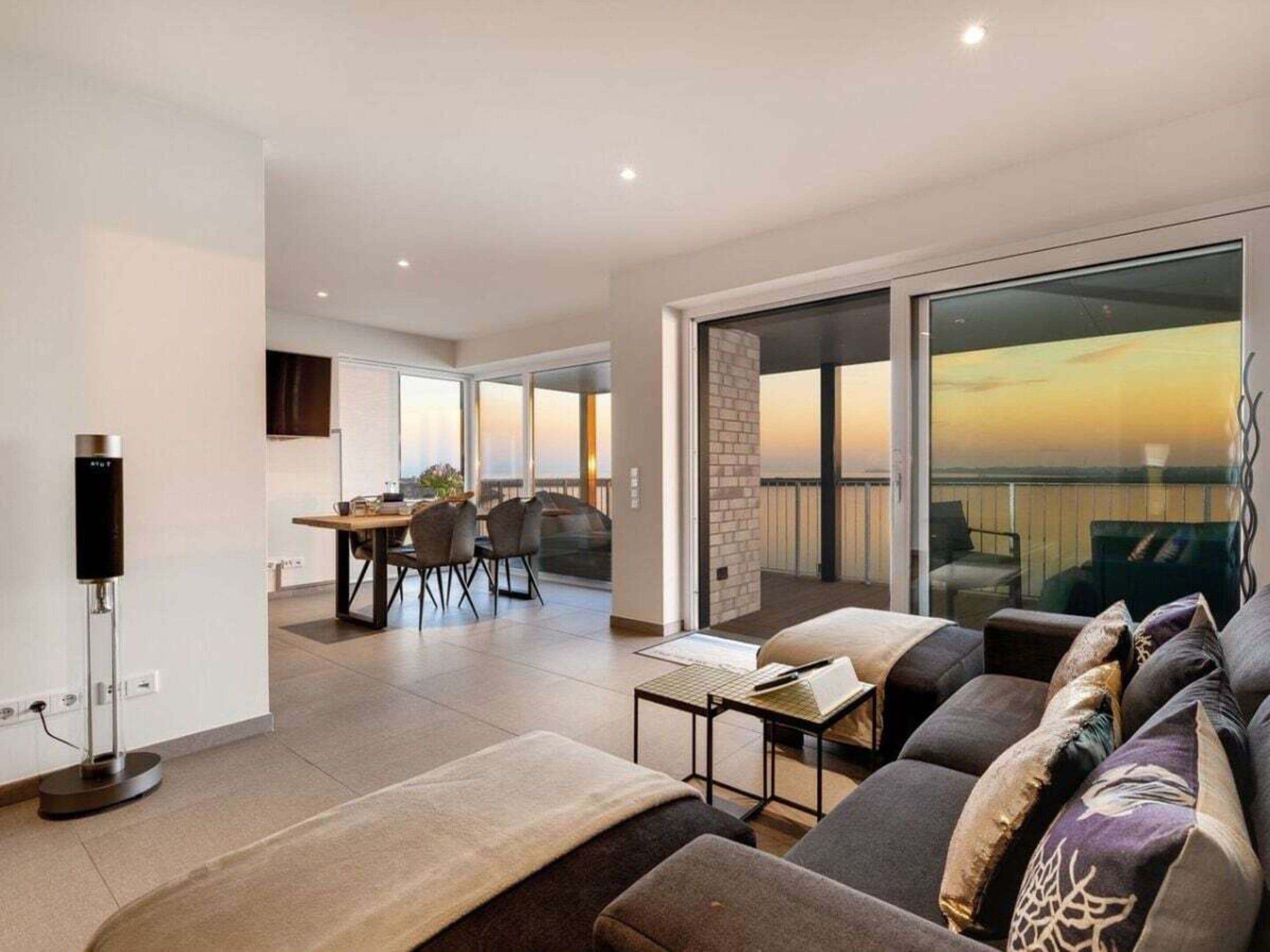 A modern living room with a sofa, a coffee table, and a dining area with chairs. There is a balcony with a view.