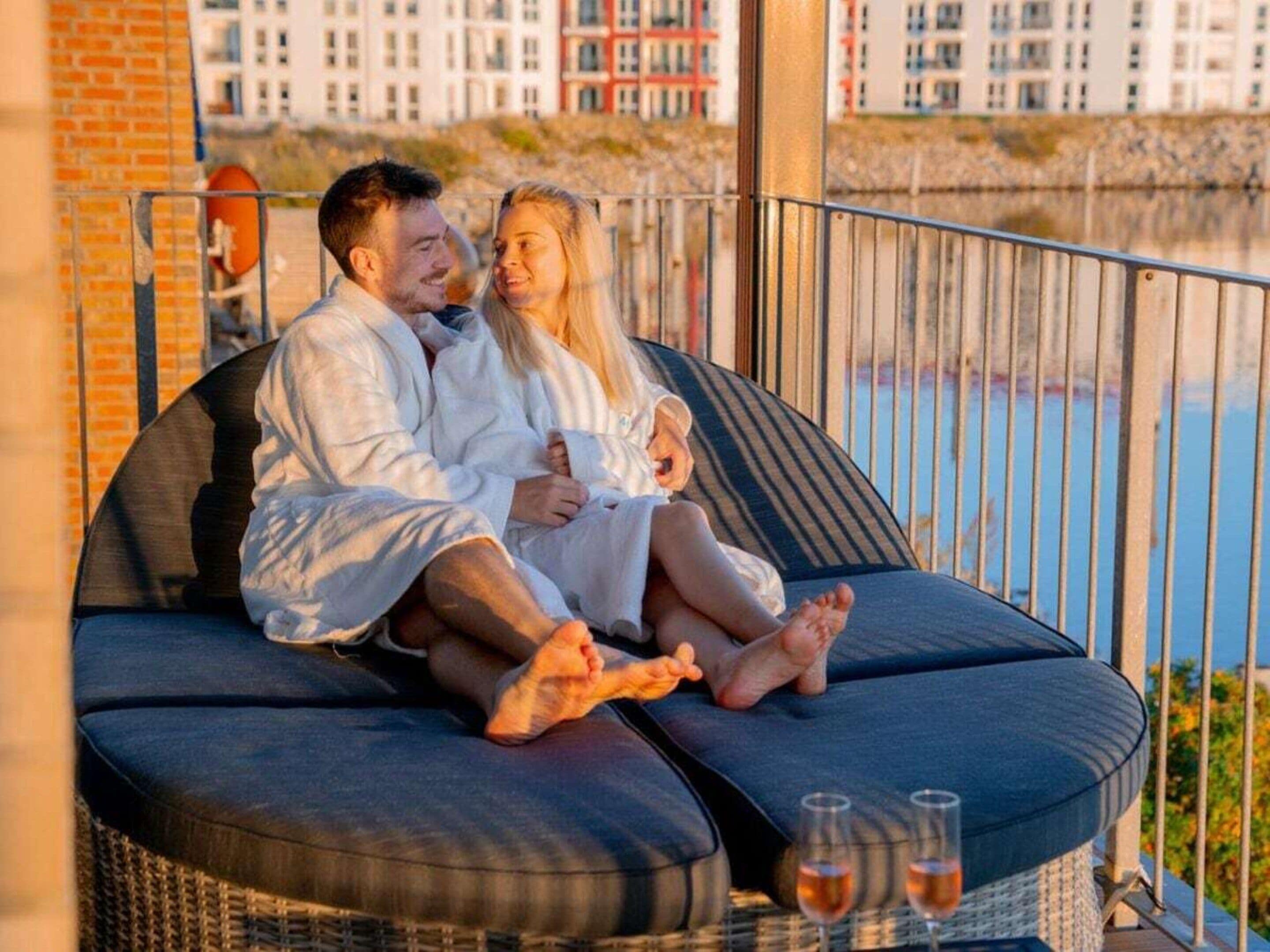 A couple sitting on a curved outdoor sofa with a view of a body of water and buildings in the background.