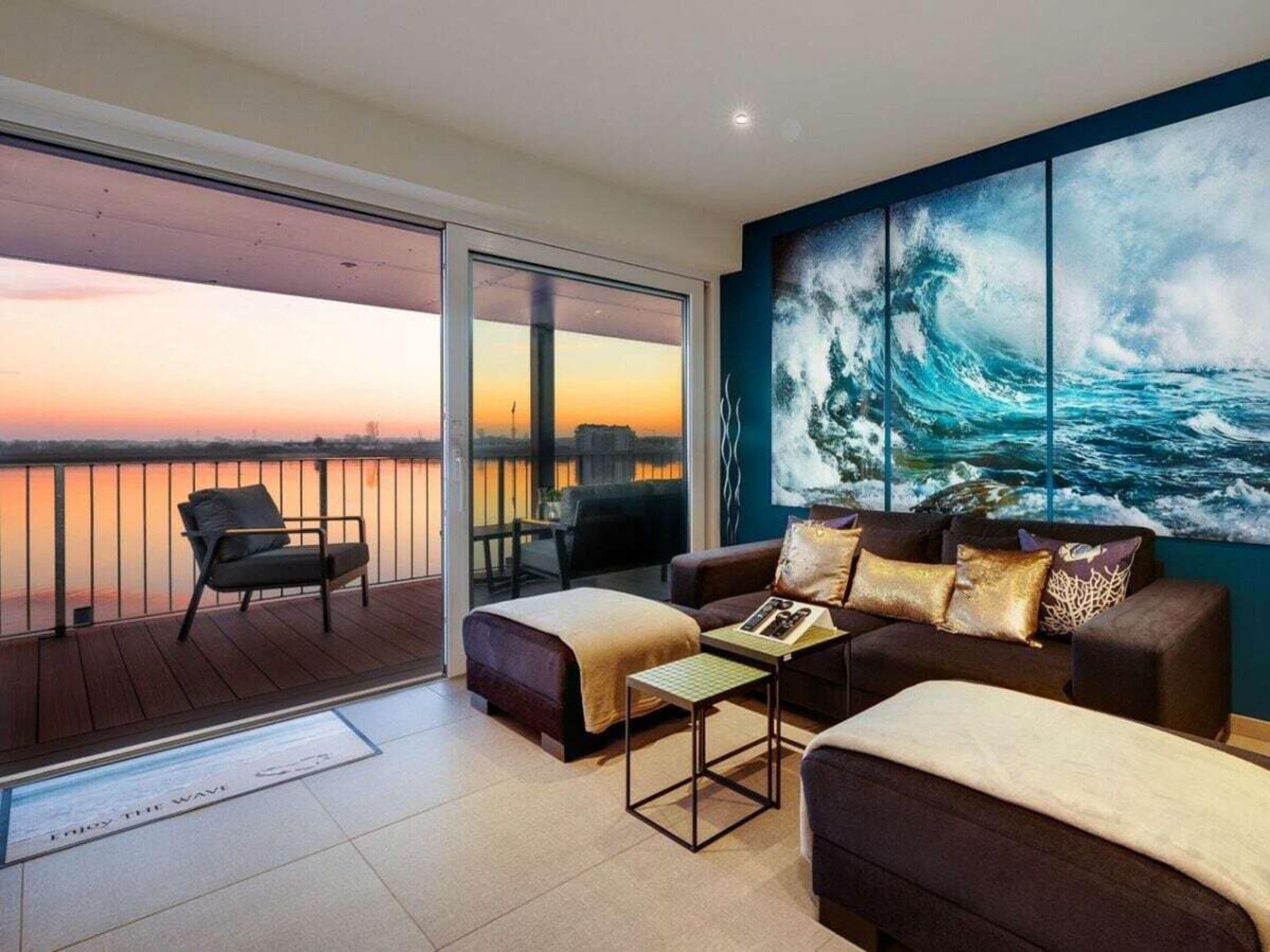 A modern living room with a balcony overlooking a body of water, featuring a large ocean wave painting on the wall.
