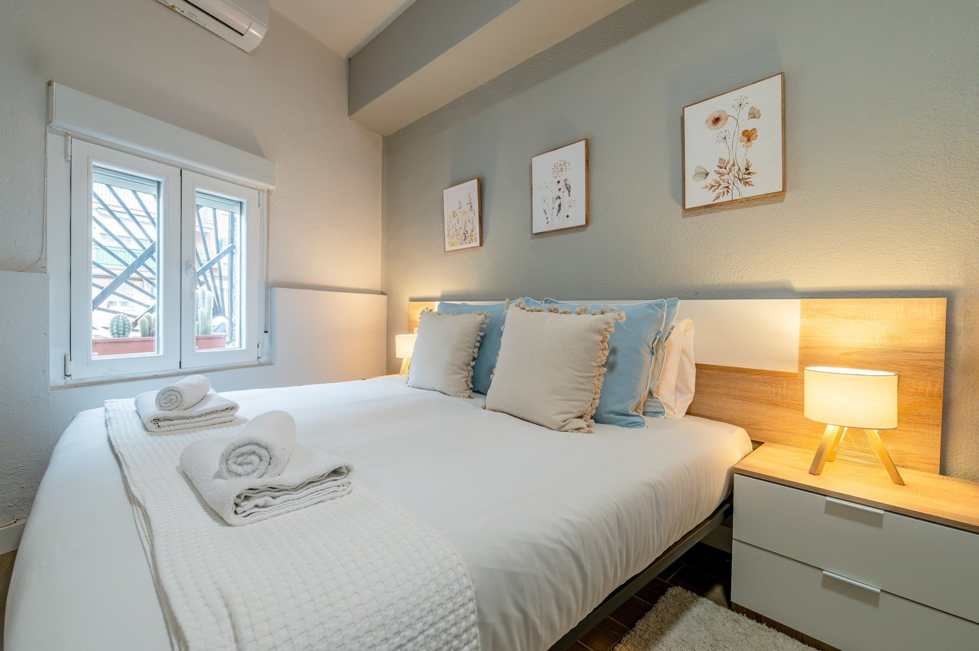 A neatly made bed with pillows and a folded towel, a bedside table with a lamp, and a window with a view of cacti.