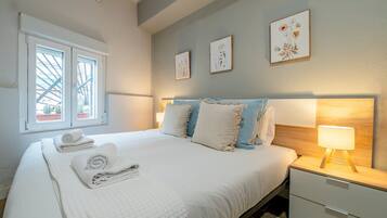A neatly made bed with pillows and a folded towel, a bedside table with a lamp, and a window with a view of cacti.