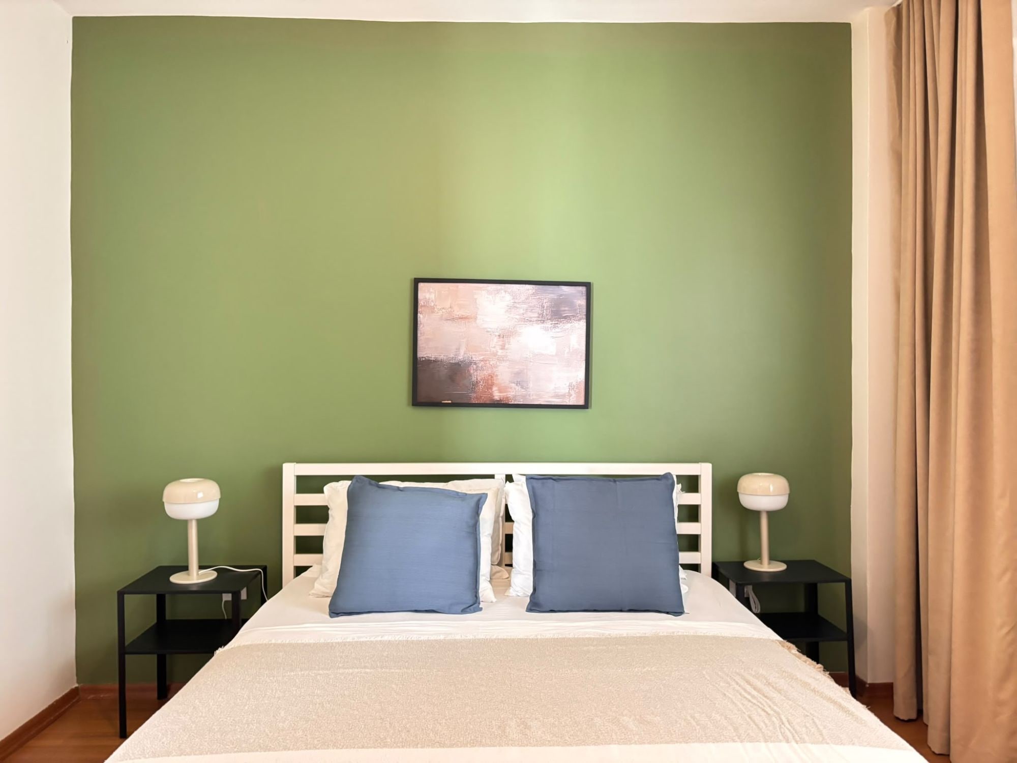 A bedroom with a bed, two bedside tables, a nightstand, a wall-mounted lamp, and a window with curtains.