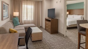 A hotel room with a sofa, a TV on a wooden stand, a small table, and a bed with white linens. - 1 Bedroom King Suite w/ Full Kitchen + Breakfast (Hillsboro)