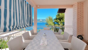Um terraço com mesa xadrez, cadeiras de vime branco, toldo listrado e vista para o mar.