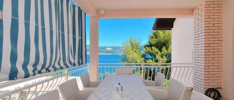 A terrace with a checkered table, white wicker chairs, a striped awning, and a view of the sea.