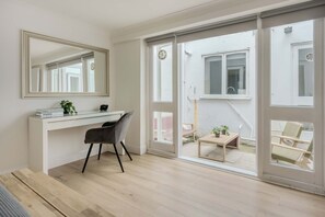 Ein modernes Zimmer mit einem weißen Schreibtisch, einem Sessel, einem großen Spiegel und einer Glastür, die zu einem Außenbereich mit Tisch und Stühlen führt.